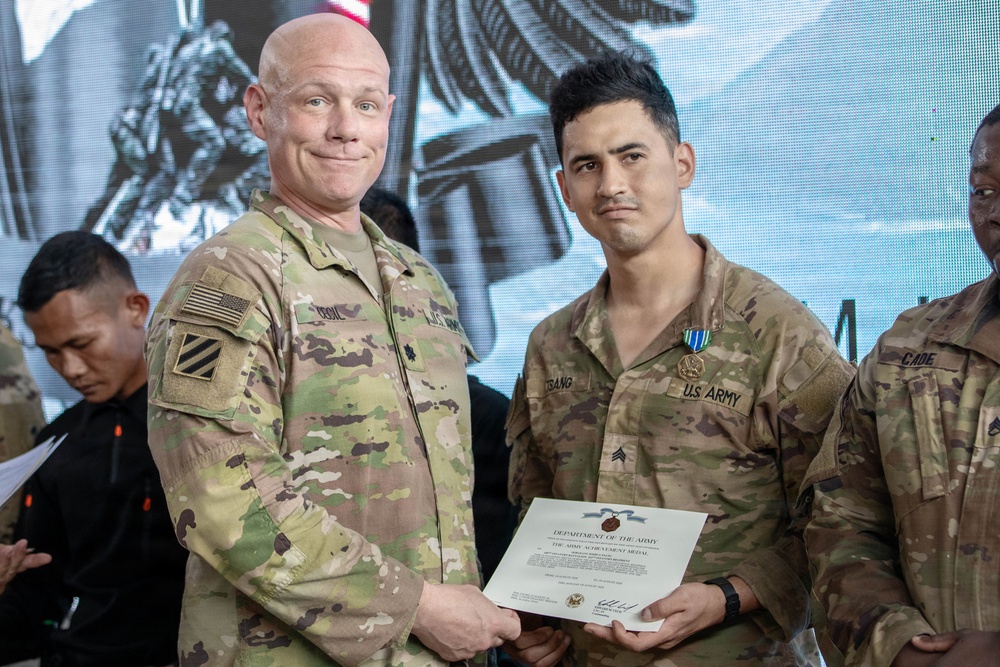 Two U.S. Army soldiers are posing together during a military awards ceremony. The soldier on the right is receiving a Department of the Army certificate for the Army Achievement Medal, which is also pinned to his uniform. A third soldier is partially visible on the right edge of the frame.