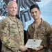 Two U.S. Army soldiers are posing together during a military awards ceremony. The soldier on the right is receiving a Department of the Army certificate for the Army Achievement Medal, which is also pinned to his uniform. A third soldier is partially visible on the right edge of the frame.