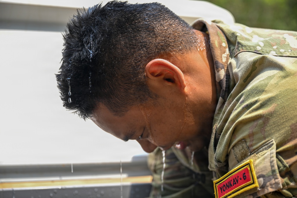 A soldier in camouflage uniform is leaning over a water source, with water visibly dripping from his wet hair and face. A name tape or unit patch reading 'YONKAV-6' is visible on his right shoulder sleeve. The image appears to capture a moment of cooling off or washing up during a military exercise or field operation.