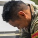 A soldier in camouflage uniform is leaning over a water source, with water visibly dripping from his wet hair and face. A name tape or unit patch reading 'YONKAV-6' is visible on his right shoulder sleeve. The image appears to capture a moment of cooling off or washing up during a military exercise or field operation.