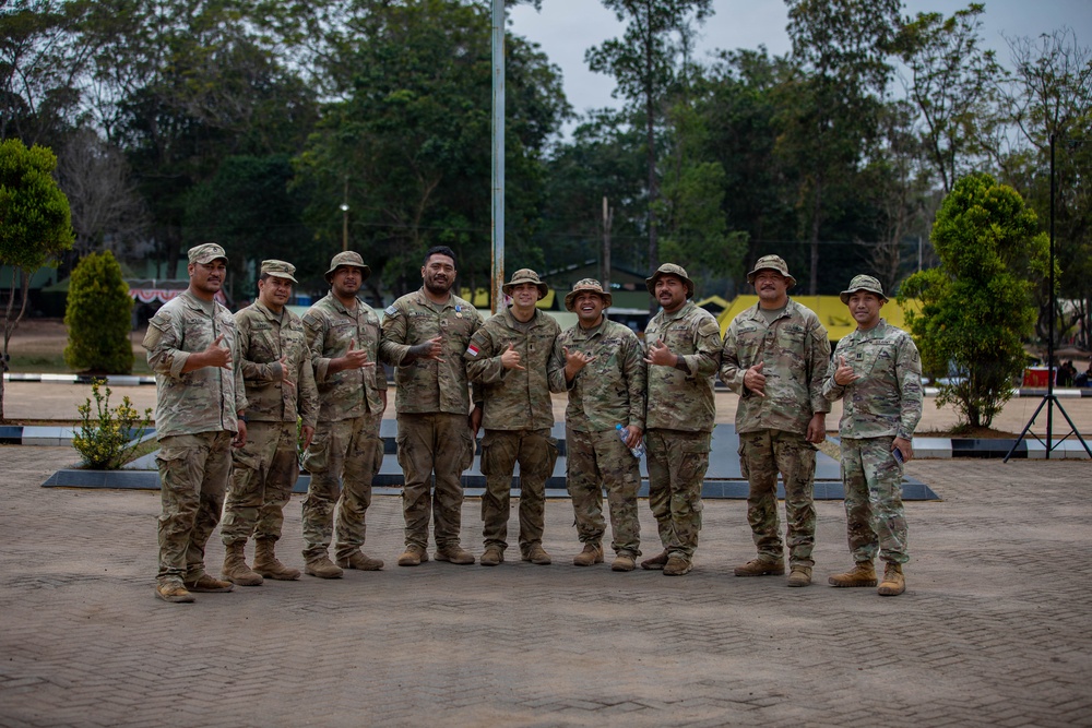 A group of nine military personnel in camouflage uniforms pose together for a group photo on a paved outdoor area. Several individuals are making 'shaka' (hang loose) hand gestures, suggesting a relaxed and celebratory atmosphere. The setting appears to be a military installation or training facility with trees, trimmed shrubs, and structures visible in the background.