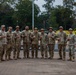 A group of nine military personnel in camouflage uniforms pose together for a group photo on a paved outdoor area. Several individuals are making 'shaka' (hang loose) hand gestures, suggesting a relaxed and celebratory atmosphere. The setting appears to be a military installation or training facility with trees, trimmed shrubs, and structures visible in the background.