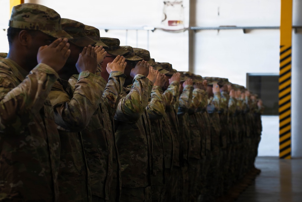 A formation of military personnel in Operational Camouflage Pattern (OCP) uniforms are rendering a salute in unison inside what appears to be a hangar or large military facility. The soldiers are aligned in a single-file row extending into the background, all performing the hand salute simultaneously. The image captures a formal military ceremony or formation event.
