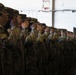 A formation of military personnel in Operational Camouflage Pattern (OCP) uniforms are rendering a salute in unison inside what appears to be a hangar or large military facility. The soldiers are aligned in a single-file row extending into the background, all performing the hand salute simultaneously. The image captures a formal military ceremony or formation event.