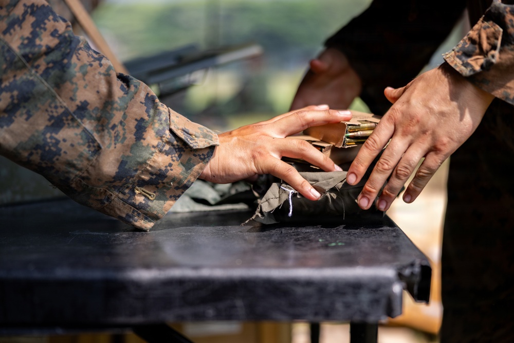 Two individuals wearing military camouflage uniforms are handling ammunition and gear on a dark surface, likely a table at a firing range or training area. Their hands are visible interacting with what appears to be rifle cartridges/rounds and a dark fabric pouch or bag. The scene suggests a military training or range preparation activity.