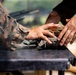 Two individuals wearing military camouflage uniforms are handling ammunition and gear on a dark surface, likely a table at a firing range or training area. Their hands are visible interacting with what appears to be rifle cartridges/rounds and a dark fabric pouch or bag. The scene suggests a military training or range preparation activity.