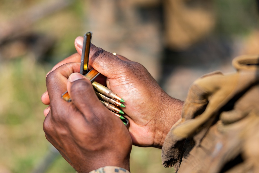 A close-up photograph shows a person in military or tactical attire handling ammunition. The individual is loading or organizing rifle cartridges with green-tipped bullets into a magazine. The shallow depth of field keeps the hands and ammunition in sharp focus while the background is blurred.