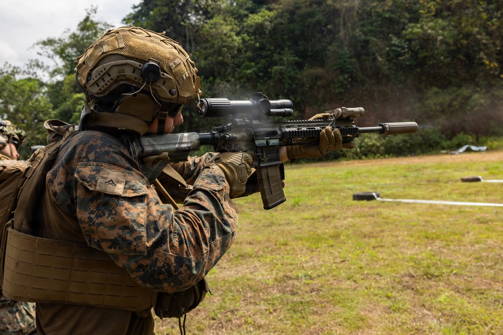 A U.S. Marine in MARPAT digital camouflage uniform is actively firing a rifle at what appears to be an outdoor shooting range. The weapon is visibly discharging, with smoke/gas visible at the muzzle and ejection port. The setting appears to be a tropical or subtropical environment based on the lush green vegetation in the background.