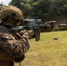 A U.S. Marine in MARPAT digital camouflage uniform is actively firing a rifle at what appears to be an outdoor shooting range. The weapon is visibly discharging, with smoke/gas visible at the muzzle and ejection port. The setting appears to be a tropical or subtropical environment based on the lush green vegetation in the background.