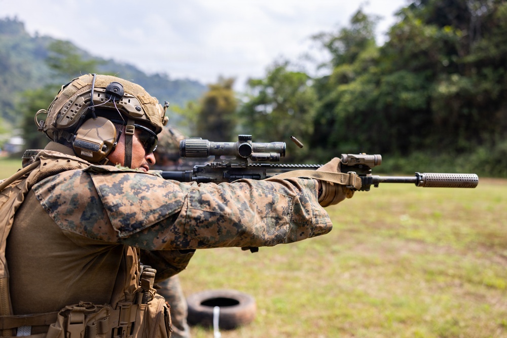A military service member in MARPAT (Marine pattern) digital camouflage is actively firing a rifle at what appears to be an outdoor shooting range. A spent brass casing is visibly ejecting from the weapon's ejection port, indicating the weapon has just been discharged. The setting appears to be a tropical or subtropical environment based on the lush green vegetation in the background.