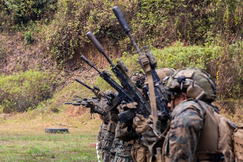 A line of military personnel in full combat gear are conducting a live-fire range exercise, aiming rifles downrange in a coordinated firing line. The soldiers are wearing woodland MARPAT camouflage uniforms consistent with U.S. Marines, equipped with tactical vests, helmets, and hearing protection. The setting appears to be an outdoor shooting range with a natural earthen berm backstop.