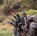 A line of military personnel in full combat gear are conducting a live-fire range exercise, aiming rifles downrange in a coordinated firing line. The soldiers are wearing woodland MARPAT camouflage uniforms consistent with U.S. Marines, equipped with tactical vests, helmets, and hearing protection. The setting appears to be an outdoor shooting range with a natural earthen berm backstop.