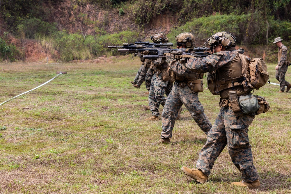 A line of military personnel in full combat gear are conducting a live-fire or tactical training exercise on an open grassy field. They are advancing in a file formation with rifles raised and aimed forward. A supervisor or range safety officer is visible walking parallel to the line on the right side.