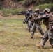 A line of military personnel in full combat gear are conducting a live-fire or tactical training exercise on an open grassy field. They are advancing in a file formation with rifles raised and aimed forward. A supervisor or range safety officer is visible walking parallel to the line on the right side.