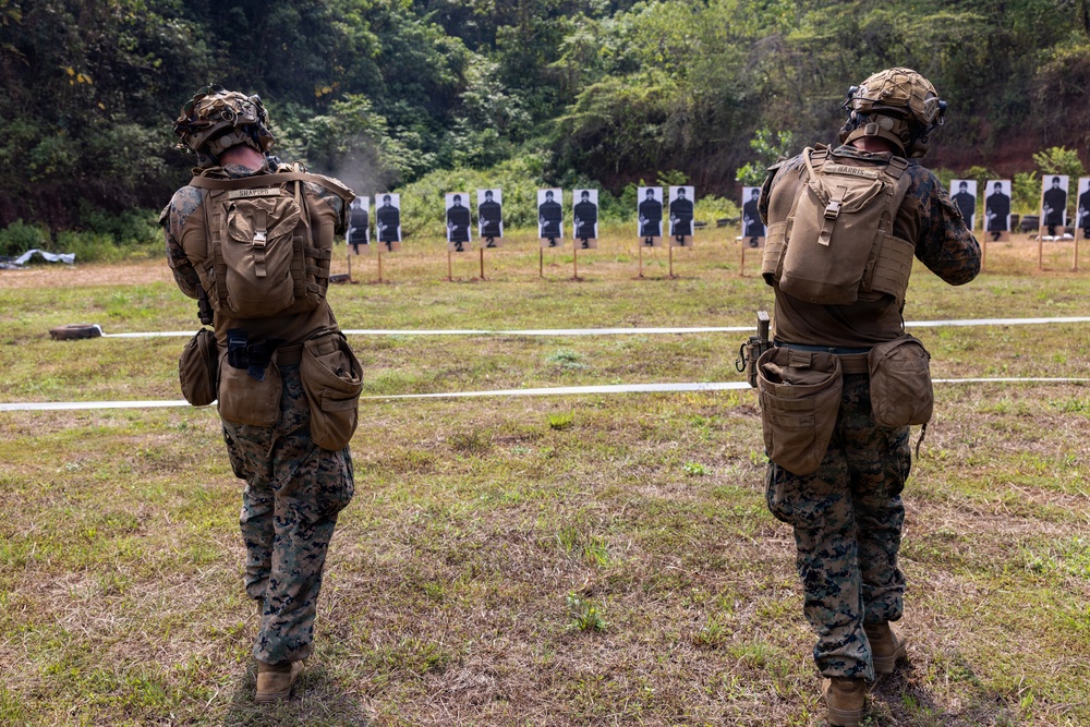 Two military personnel in full combat gear stand at a firing range facing a row of silhouette targets downrange. Both individuals are wearing USMC MARPAT camouflage uniforms with tactical vests, helmets, and backpacks. The setting appears to be an outdoor range bordered by dense vegetation.