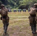 Two military personnel in full combat gear stand at a firing range facing a row of silhouette targets downrange. Both individuals are wearing USMC MARPAT camouflage uniforms with tactical vests, helmets, and backpacks. The setting appears to be an outdoor range bordered by dense vegetation.