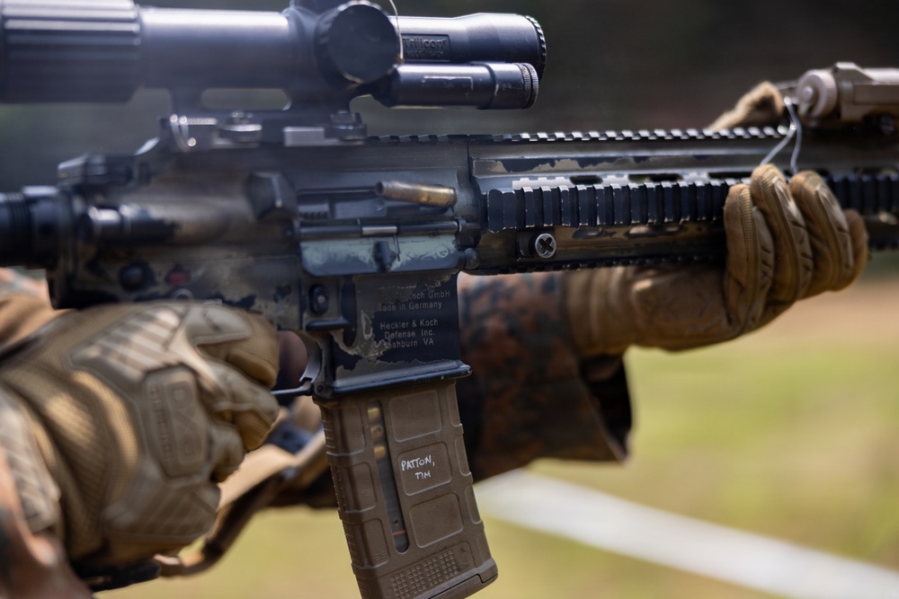 A close-up photograph of a Hecker &amp; Koch rifle being held by a person wearing tactical gloves in a camouflage pattern. The weapon is equipped with a rifle scope, Picatinny rail system, and a tan MAGPUL magazine marked with the name 'PATTON, TIM'. The image appears to be taken at an outdoor shooting range or training environment.