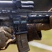 A close-up photograph of a Hecker &amp; Koch rifle being held by a person wearing tactical gloves in a camouflage pattern. The weapon is equipped with a rifle scope, Picatinny rail system, and a tan MAGPUL magazine marked with the name 'PATTON, TIM'. The image appears to be taken at an outdoor shooting range or training environment.