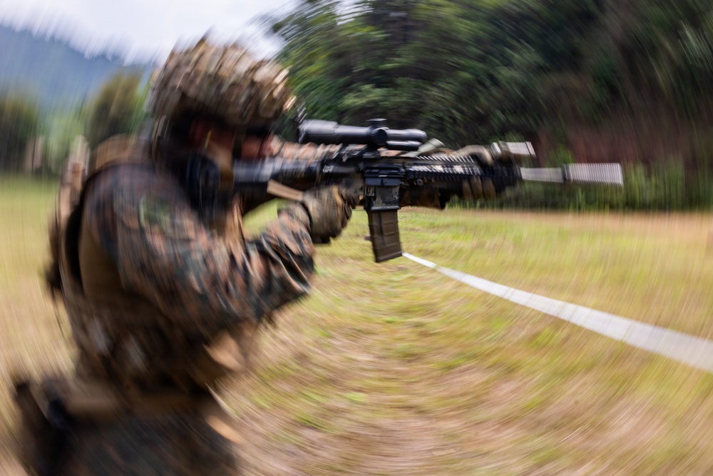 A soldier in camouflage uniform and helmet is aiming a rifle with an optical scope attachment, captured with a radial zoom blur photographic technique that creates a dynamic tunnel-effect composition. The image appears to be taken during a military training exercise in an outdoor field environment. The intentional camera motion blur keeps the weapon barrel as the sharpest focal point of the image.
