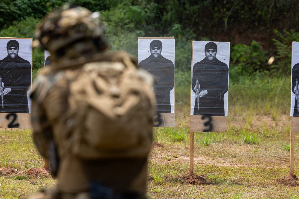 A military or law enforcement personnel in full tactical gear, including a camouflage helmet and body armor, faces a row of paper shooting targets at an outdoor firing range. The targets display a printed human silhouette figure and are numbered, with at least targets 2 and 3 visible. The setting appears to be a professional training exercise conducted in a cleared outdoor area with dense vegetation in the background.
