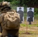 A military or law enforcement personnel in full tactical gear, including a camouflage helmet and body armor, faces a row of paper shooting targets at an outdoor firing range. The targets display a printed human silhouette figure and are numbered, with at least targets 2 and 3 visible. The setting appears to be a professional training exercise conducted in a cleared outdoor area with dense vegetation in the background.