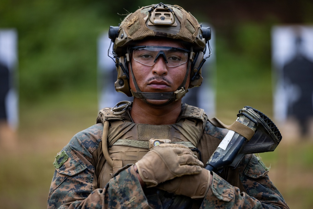 A U.S. Marine in full combat gear stands at what appears to be a shooting range, holding a firearm at a low-ready position. The Marine is equipped with tactical helmet, hearing protection, eye protection, body armor, and digital camouflage uniform. Paper target silhouettes are visible in the blurred background.