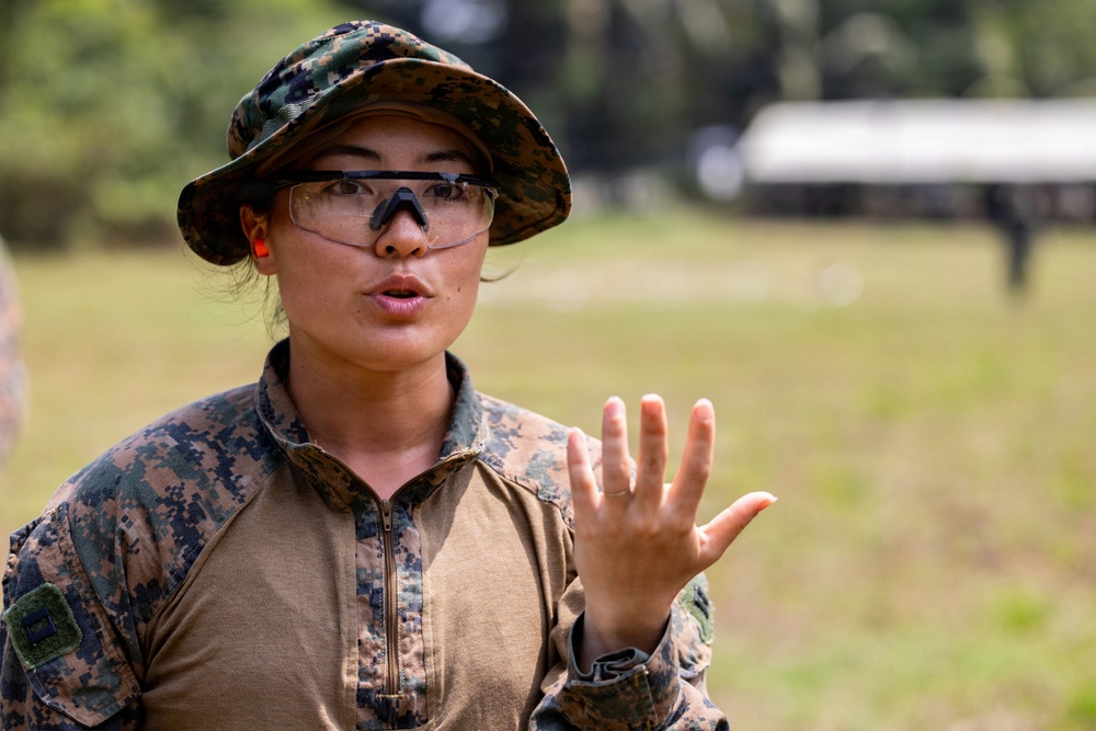 A female military service member wearing MARPAT (Marine pattern) digital camouflage uniform and a boonie cover is speaking or giving instruction outdoors. She is wearing protective eyewear and orange ear protection, and is gesturing with her right hand raised, fingers spread. The background shows a grassy field with trees and a structure, consistent with a military training range environment.