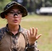 A female military service member wearing MARPAT (Marine pattern) digital camouflage uniform and a boonie cover is speaking or giving instruction outdoors. She is wearing protective eyewear and orange ear protection, and is gesturing with her right hand raised, fingers spread. The background shows a grassy field with trees and a structure, consistent with a military training range environment.