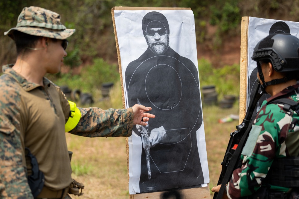 Two military personnel are examining a paper shooting target at an outdoor firing range. One individual wearing US Marine Corps MARPAT camouflage and a patrol cap is pointing at bullet impact locations on the target. The other individual is wearing green camouflage and a tactical helmet, consistent with a foreign military partner force.