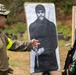 Two military personnel are examining a paper shooting target at an outdoor firing range. One individual wearing US Marine Corps MARPAT camouflage and a patrol cap is pointing at bullet impact locations on the target. The other individual is wearing green camouflage and a tactical helmet, consistent with a foreign military partner force.