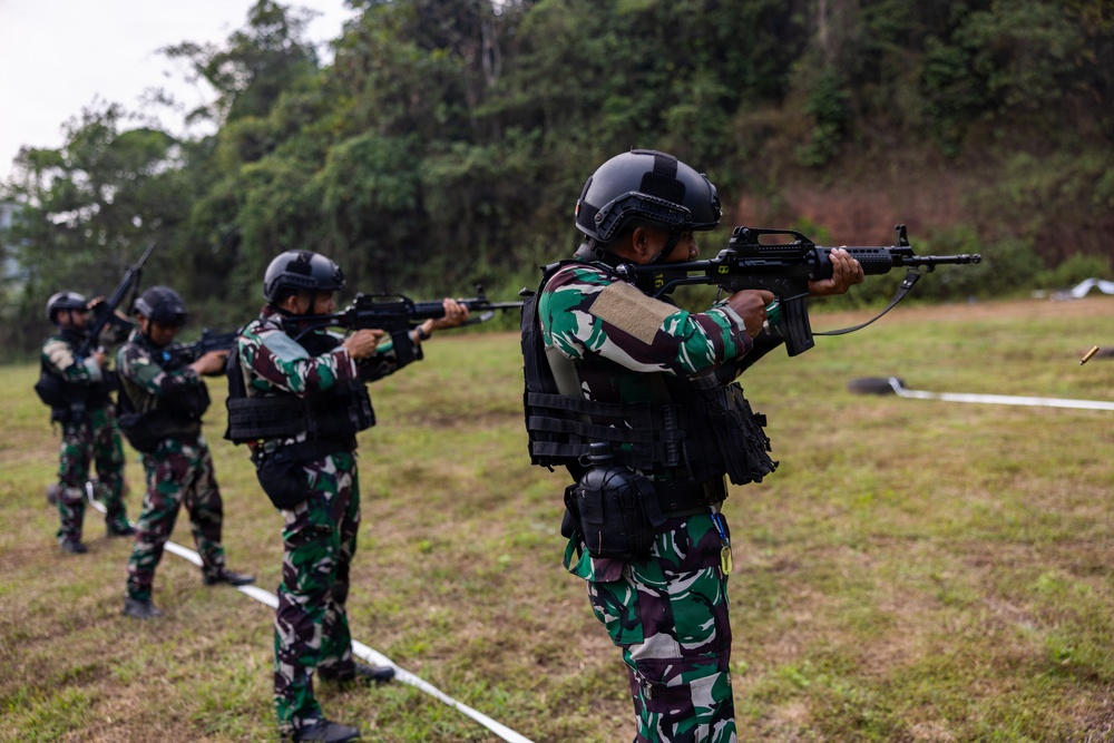 A group of military or special forces personnel are conducting a live-fire shooting exercise on an outdoor range. The soldiers are wearing matching camouflage uniforms with tactical vests and black helmets, firing rifles in a line formation. A spent brass casing is visibly ejecting from the nearest shooter's weapon, confirming active firing.