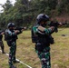 A group of military or special forces personnel are conducting a live-fire shooting exercise on an outdoor range. The soldiers are wearing matching camouflage uniforms with tactical vests and black helmets, firing rifles in a line formation. A spent brass casing is visibly ejecting from the nearest shooter's weapon, confirming active firing.