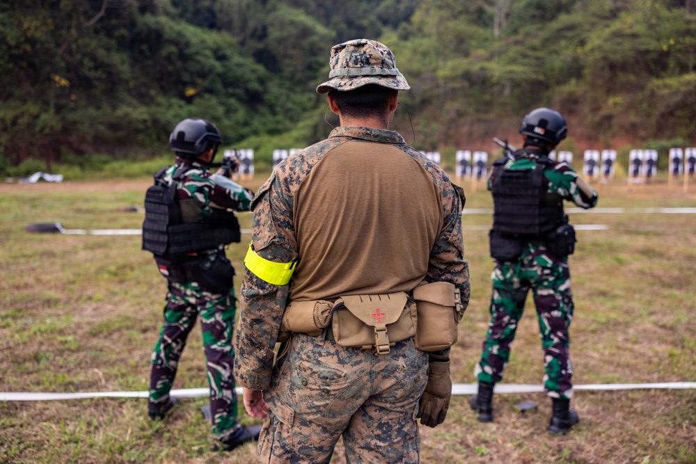 A military shooting range training exercise is depicted, showing three personnel facing downrange toward multiple paper silhouette targets. The central figure, wearing US Marine Corps MARPAT digital camouflage with a yellow safety armband and a medical pouch, appears to be an instructor overseeing two other soldiers in a different camouflage pattern. The setting is an outdoor range bordered by dense tropical vegetation.