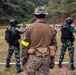 A military shooting range training exercise is depicted, showing three personnel facing downrange toward multiple paper silhouette targets. The central figure, wearing US Marine Corps MARPAT digital camouflage with a yellow safety armband and a medical pouch, appears to be an instructor overseeing two other soldiers in a different camouflage pattern. The setting is an outdoor range bordered by dense tropical vegetation.