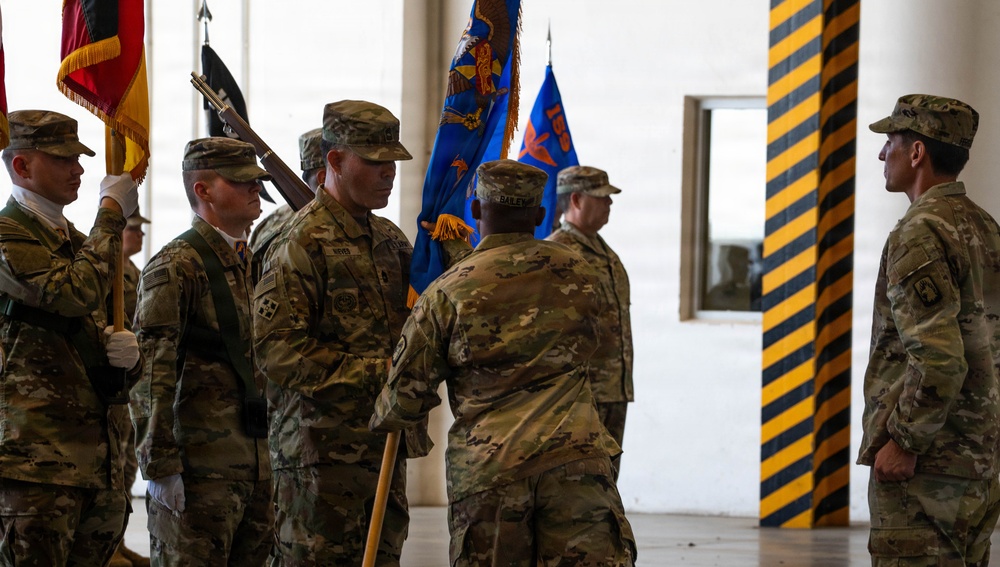 A military ceremony is taking place inside what appears to be a large indoor facility, likely a hangar or drill hall. Soldiers in U.S. Army OCP (Operational Camouflage Pattern) uniforms are participating in what appears to be a change of command or unit activation/deactivation ceremony, evidenced by the passing of unit colors. Multiple flags and guidons are visible, including what appears to be a German flag and blue unit colors.
