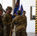 A military ceremony is taking place inside what appears to be a large indoor facility, likely a hangar or drill hall. Soldiers in U.S. Army OCP (Operational Camouflage Pattern) uniforms are participating in what appears to be a change of command or unit activation/deactivation ceremony, evidenced by the passing of unit colors. Multiple flags and guidons are visible, including what appears to be a German flag and blue unit colors.