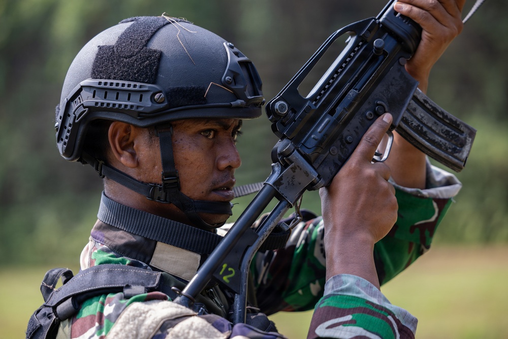 A soldier wearing a black tactical helmet and camouflage uniform is inspecting or handling a firearm at close range. The individual appears to be examining the weapon carefully, possibly performing a function check or clearing procedure. The background shows a blurred green outdoor environment consistent with a training or field setting.