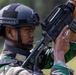 A soldier wearing a black tactical helmet and camouflage uniform is inspecting or handling a firearm at close range. The individual appears to be examining the weapon carefully, possibly performing a function check or clearing procedure. The background shows a blurred green outdoor environment consistent with a training or field setting.
