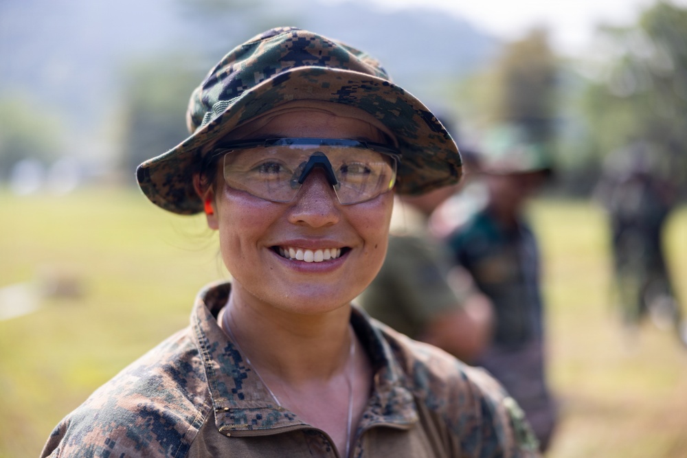 A smiling individual wearing digital camouflage uniform and boonie hat with clear protective eyewear is photographed in an outdoor field setting. The subject appears to be part of a military or paramilitary group, with additional uniformed personnel visible in the blurred background. The image conveys a positive, candid moment during what appears to be a field exercise or training activity.