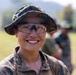 A smiling individual wearing digital camouflage uniform and boonie hat with clear protective eyewear is photographed in an outdoor field setting. The subject appears to be part of a military or paramilitary group, with additional uniformed personnel visible in the blurred background. The image conveys a positive, candid moment during what appears to be a field exercise or training activity.