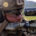 A close-up photograph of a military service member in full tactical gear aiming down a rifle with an optical scope. The soldier is wearing a combat helmet with mounted accessories, tactical eye protection, and hearing protection. The background shows a blurred outdoor environment with green terrain and hills.