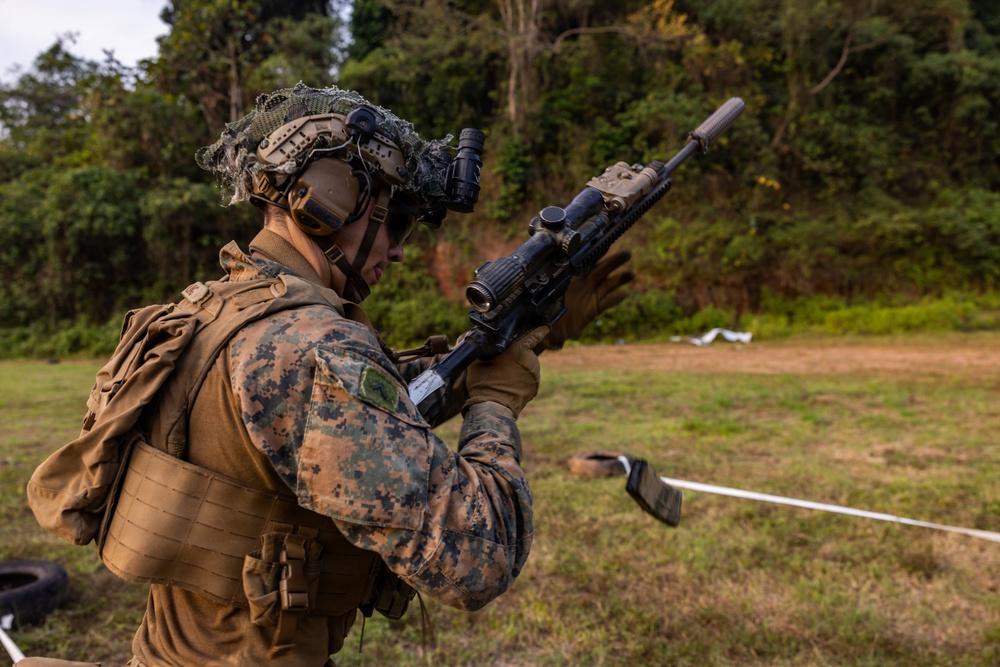 A military service member in MARPAT (Marine pattern) camouflage uniform is holding a rifle with a suppressor and optical scope at a training range. The soldier is fully equipped with tactical gear including a helmet with night vision mount, body armor, and a backpack. The setting appears to be an outdoor field training environment with a grassy clearing and tree line in the background.