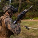 A military service member in MARPAT (Marine pattern) camouflage uniform is holding a rifle with a suppressor and optical scope at a training range. The soldier is fully equipped with tactical gear including a helmet with night vision mount, body armor, and a backpack. The setting appears to be an outdoor field training environment with a grassy clearing and tree line in the background.