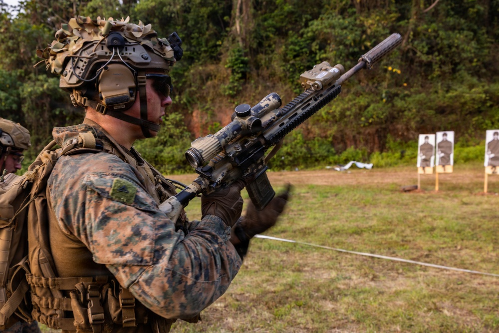 A heavily equipped military service member in MARPAT camouflage uniform is holding an assault rifle with a suppressor and optic scope at a live-fire shooting range. The soldier is wearing advanced combat gear including a tactical helmet with night vision mounts, communications headset, and body armor. Silhouette paper targets are visible in the background, indicating an active training exercise.