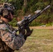 A heavily equipped military service member in MARPAT camouflage uniform is holding an assault rifle with a suppressor and optic scope at a live-fire shooting range. The soldier is wearing advanced combat gear including a tactical helmet with night vision mounts, communications headset, and body armor. Silhouette paper targets are visible in the background, indicating an active training exercise.