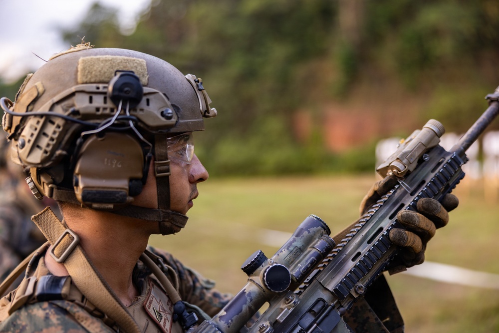 A military service member in full tactical gear is holding and inspecting a rifle equipped with optical attachments. The subject appears to be at an outdoor training range or field exercise, wearing MARPAT camouflage uniform consistent with U.S. Marine Corps. The image is a close-up profile shot with a shallow depth of field, keeping the background blurred.
