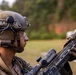 A military service member in full tactical gear is holding and inspecting a rifle equipped with optical attachments. The subject appears to be at an outdoor training range or field exercise, wearing MARPAT camouflage uniform consistent with U.S. Marine Corps. The image is a close-up profile shot with a shallow depth of field, keeping the background blurred.