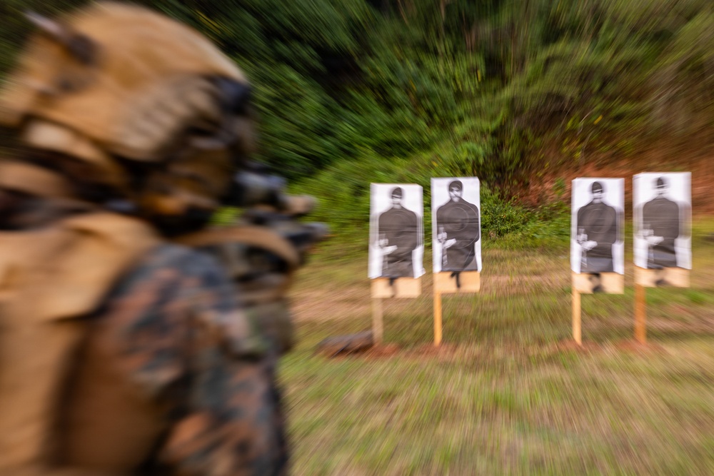 A military or law enforcement personnel in camouflage uniform and tactical gear is engaged in a shooting range exercise. Four paper silhouette targets are mounted on wooden stakes downrange in an outdoor wooded setting. The image uses a zoom-burst photographic technique, creating motion blur radiating from the targets.