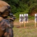 A military or law enforcement personnel in camouflage uniform and tactical gear is engaged in a shooting range exercise. Four paper silhouette targets are mounted on wooden stakes downrange in an outdoor wooded setting. The image uses a zoom-burst photographic technique, creating motion blur radiating from the targets.