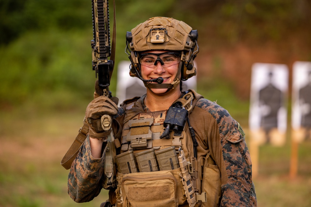 A smiling military service member in full combat gear stands at what appears to be a shooting range. The individual is wearing MARPAT (Marine pattern) camouflage uniform, a tactical helmet with mounted equipment, and a plate carrier vest, holding a rifle upright. Shooting targets are visible in the blurred background.